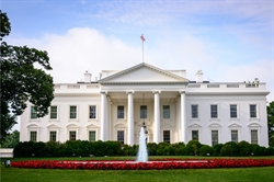 [ai] The White House, a historic white mansion with large columns, a green lawn, and red flower beds in the foreground. A fountain is visible in front of the building, under a blue sky with white clouds.